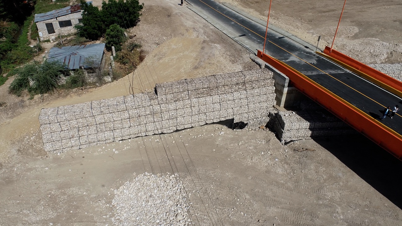 Vista aérea de un muro de gaviones junto al acceso a un puente con barandas anaranjadas.